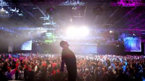 Tony Robbins no palco, contra a luz. Uma multidão lota uma arena multiuso para ver suas palestras. Desperte seu gigante interior 